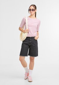 Gestreiftes rosa und weißes figurbetontes Oberteil, dunkle Denim-Bermuda-Shorts mit fransigem Saum, rosa Sneaker mit orangefarbenen Streifen und eine hellfarbene Schultertasche.