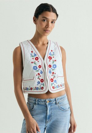 Joven mujer que lleva un chaleco blanco acolchado sin mangas con bordados florales rojos y azules y vaqueros azul claro, de pie frente a un fondo liso.