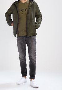 Oliven grøn zip-up jakke med hætte, olivengrøn t-shirt med sort tekst, mørkegrå jeans og hvide sneakers. Glat materiale hele vejen igennem.