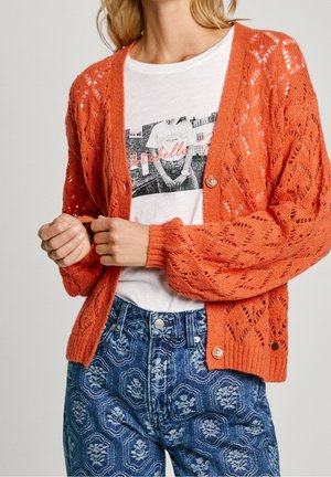 Vrouw draagt een oranje gebreid vest over een wit t-shirt met print en blauwe, hoog getailleerde jeans met patroon, en past de mouw van het vest aan.