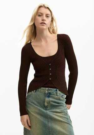 Blondína v priliehavom tmavohnedom rebrovanom kardigane s gombíkmi cez zodpovedajúci top a vyblednutú modrú denimovú sukňu.