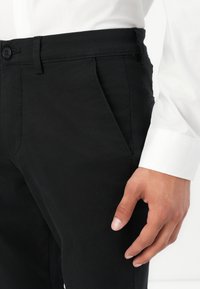 Schwarze, maßgeschneiderte Hosen mit glatter Textur, ausgestattet mit seitlichen Taschen, einem Knopfverschluss und einem schmalen Design, kombiniert mit einem weißen Hemd.