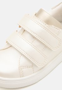 Baskets blanches avec une texture en cuir lisse, dotées de deux bandoulières Velcro et d'un accent doré sur le col rembourré. Semelle en caoutchouc avec une adhérence texturée.
