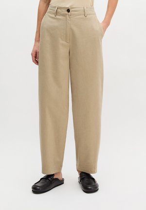 Pantalon beige taille haute à jambes larges avec poches latérales, porté avec des mocassins en cuir noir à enfiler, sur fond blanc.