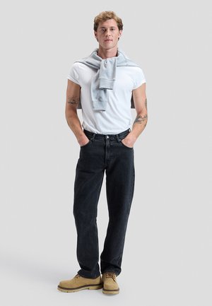 Jeune homme avec des tatouages portant un t-shirt blanc, un pull gris sur les épaules, un jean noir et des bottes beige, debout avec les mains dans les poches.