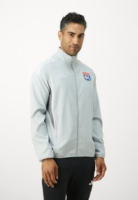 Hellgraue Sportjacke mit hohem Kragen, Reißverschluss und Adidas-Logo. Sie verfügt über drei weiße Streifen an den Ärmeln und ein Teamabzeichen.