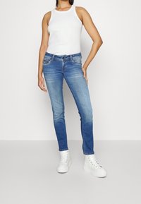 Jeans ajustados de color azul con un ligero desgastado, cinco bolsillos y acentos metálicos, combinados con una camiseta sin mangas blanca y zapatillas altas blancas.