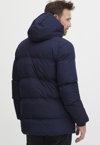 Mørkeblå dunjakke med hette, med en quiltet design og elastiske mansjetter. Bakviewet viser fremtredende sømmer og en avslappet passform.