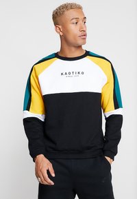 Sweatshirt preto com mangas raglan multicoloridas em amarelo, verde-água e branco. Apresenta um logótipo em texto preto. Feito de tecido macio.