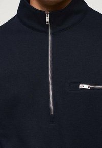 Pullover blu navy con cerniera frontale e collo alto, dotato di zip argentata sul davanti e una piccola tasca sul petto con zip. Tessuto morbido e strutturato.