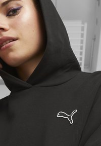 Čierna mikina s kapucňou vyrobená z mäkkej tkaniny. Obsahuje biele vyšívané logo Puma na hrudi, s hladkým, beztextúrovaným povrchom.