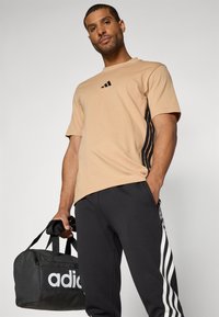 Beige bomulds T-shirt med sort logo, parret med sorte sweatsbukser med hvide vertikale striber. Holder en sort sportstaske.