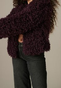 Persona che indossa un giacca testurizzata ricciola di un viola scuro e jeans neri a vita alta, tagliati in vita, davanti a uno sfondo semplice.