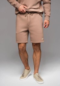 Pantaloni corti in cotone marrone con vita elasticizzata e cordoncino. Presentano dettagli di cucitura e un piccolo etichetta del marchio sul lato.