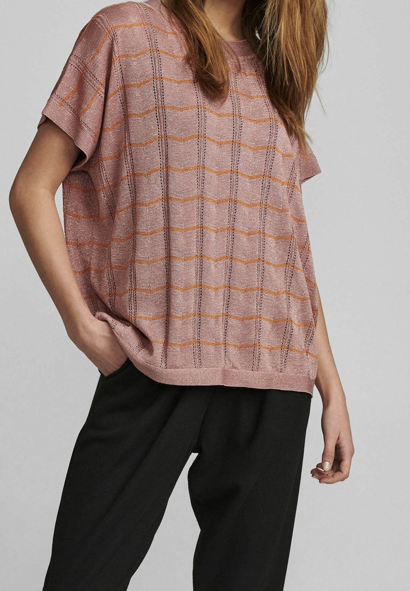 Femme portant un haut en maille rose ample à manches courtes avec des rayures horizontales orange et un pantalon noir, main dans la poche sur fond uni.