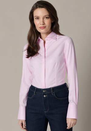 Kobieta z długimi brązowymi włosami, ubrana w jasno różową koszulę na guziki i ciemnoniebieskie dżinsy, stojąca na jednolitym beżowym tle.