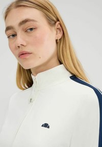 Młoda kobieta z blond włosami nosi biały dres z zamkiem, z granatowym paskiem na ramieniu i małym czarnym logo na piersi.