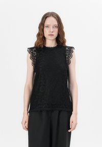 Svart blonde topp med scallop-detaljer på skuldrene, teksturert blomstermønster og rund halsringning. Båret sammen med løse svarte bukser.