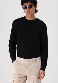 Sort ribbet sweater med lange ærmer, rund hals, bæres med beige fløjlsbukser. Modellen bærer også sorte solbriller.