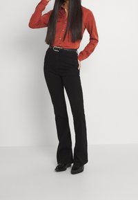 Camicia button-up color ruggine, jeans neri a palazzo, cintura nera con hardware argentato e stivali neri con tacco. Tessuto liscio e vestibilità su misura.