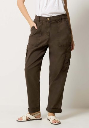 Person wearing dark brown cargo pants with large front pockets and cuffed hems paired with white crisscross sandals and a white shirt.