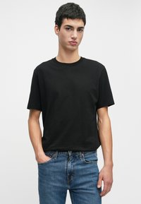 Jovem com cabelo curto e escuro, vestindo uma t-shirt preta simples e jeans azuis, de pé com uma mão no bolso contra um fundo branco.