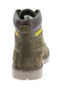 Botte en daim vert olive avec des accents en cuir marron, logo jaune, cheville rembourrée et semelle robuste. Coutures visibles le long des côtés.