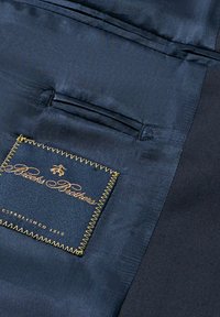 Granatowy materiał o gładkiej teksturze, z etykietą wyszywaną złotą nitką "Brooks Brothers" oraz małym detalem kieszeni nad etykietą.