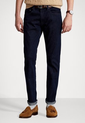 Mand iført mørkeblå opsmøgede jeans, lysegrå sokker, brune loafers med kvaster, beige sweater og et armbåndsur, der står på en hvid gulv.