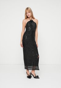 Vestido de halter negro con lentejuelas, con una silueta ajustada, abertura lateral y detalle de encaje en el dobladillo. Llevado con zapatos de tacón negros adornados con un lazo.