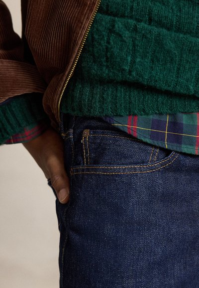 Persona que lleva vaqueros azul oscuro, suéter verde de punto sobre camisa de cuadros y chaqueta marrón de pana con cremallera, mano en el bolsillo.