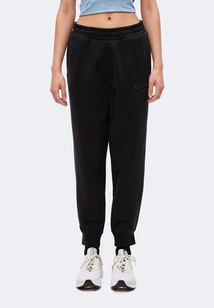 Personne portant un pantalon de jogging Puma noir avec des poignets élastiques, un top court bleu clair, des chaussettes noires et des baskets blanches, debout devant un fond blanc.