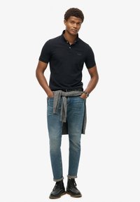 Donker marineblauwe polo met kraag, gecombineerd met lichtblauwe tapered jeans. Grijze trui om de taille geknoopt, zwarte veterschoenen.
