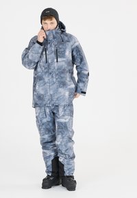 Vedeněodolná sněhová bunda a kalhoty v modro-šedé barvě s vzorem připomínajícím mraky. Obsahuje kapsy, nastavitelnou kapucu a vyztužené manžety. Černé boty.