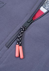 Grijze jack met rits, voorzien van een zwart-wit paracord ritstrekkertje met oranje uiteinden, die de gladde stofstructuur en subtiele stikseldetails laat zien.