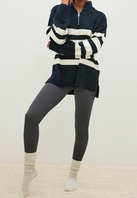 Pull rayé bleu marine et blanc avec un col zippé, associé à des leggings gris côtelés et des chaussettes en maille gris clair sur un fond crème texturé.
