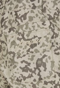 Beige cargoshorts med et struktureret, camouflage mønster i grå og grønne nuancer. Har en broderet "peqius" label og tryklås.