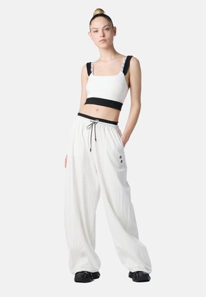 Młoda kobieta w białym krótkim topie z czarnymi ramiączkami i luźnych białych spodniach ze ściągaczem, stojąca z jedną ręką w kieszeni i w czarnych butach.