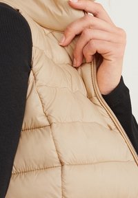 Beżowa kamizelka puchowa z wysokim kołnierzem, o pikowanej teksturze i przednim zamkiem błyskawicznym. Zbliżenie na rękę trzymającą zamek.