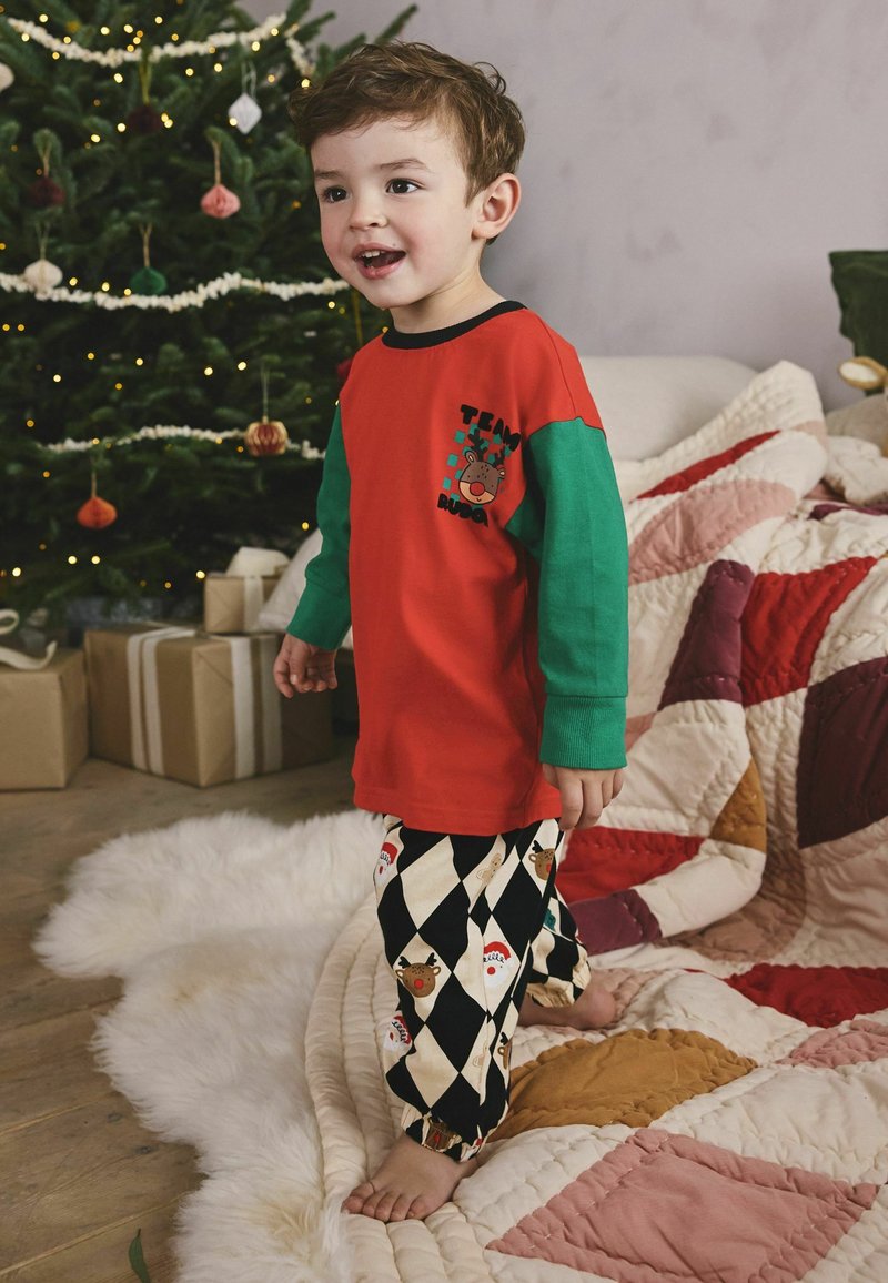 Cămașă roșie cu mâneci lungi verzi, având un grafic cu reni, asortată cu pantaloni în carouri negri și crem cu motive festive.