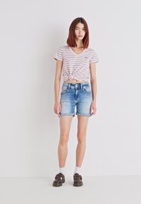 Striped, knotted pink T-shirt paired with light blue denim shorts. Model wears dark shoes and white socks, standing against a plain background.