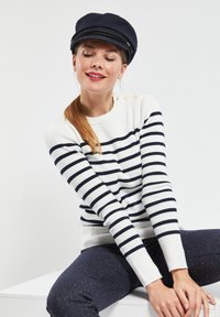 Pull blanc avec des rayures bleu marine, encolure ronde et accents de boutons sur l'épaule. Pantalons texturés bleu marine avec des fils scintillants. Casquette bleu marine.