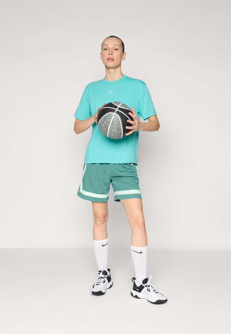 Türkisfarbiges Kurzarm-T-Shirt mit Logo, grüne Basketballshorts mit weißen Akzenten, weiße Socken und schwarz-weiße Sneaker, hält einen Basketball.