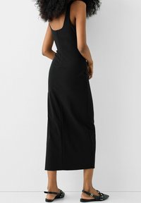 Vestito nero senza maniche con corpetto aderente e lunghezza midi. Presenta una schiena bassa e una zip nascosta. Abbinato a scarpe nere flat.