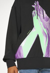Sweat à capuche noir avec un grand motif de deux mains jointes en vert et violet sur le devant, porté avec un jean bleu.