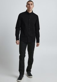 Black button-up shirt, long sleeves, solid color, paired with black pants and black sneakers, standing against a light background.