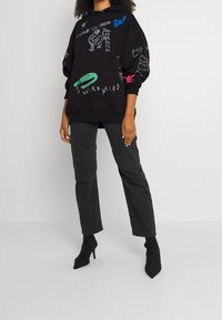 Persona con una sudadera negra de gran tamaño adornada con garabatos blancos y coloridos, y pantalones cargo rectos negros, combinados con botas de tacón negras.