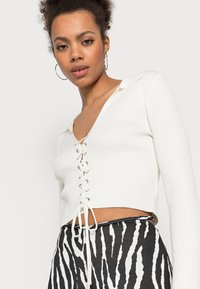 Top crop em tecido canelado branco com mangas longas, decote em V profundo e detalhes de amarração na parte da frente. Usado com calças listradas em preto e branco.