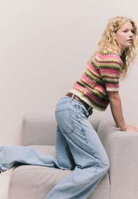 Csíkos kötött pulóver rózsaszín, zöld és fehér színekben; világoskék, bő szárú farmer; a modell egy világosszürke kanapén pózol.