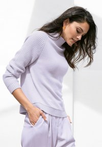 Helllila gestrickter Pullover mit einem hohen, gerippten Halsausschnitt und langen Ärmeln, kombiniert mit helllila Hosen, die eine lockere Passform und Seitentaschen aufweisen.